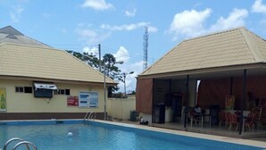 A swimming pool with a tiled deck, a covered seating area with chairs, and a building with a signboard. - Kim Royal Hotel and Suites (Asaba)