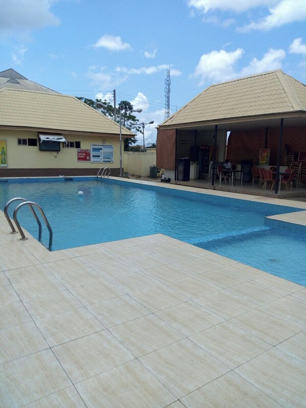 Una piscina con solárium empedrado, una zona de estar cubierta con sillas y un edificio con cartel.