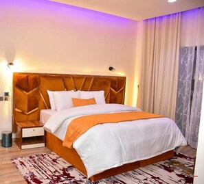Una habitación de hotel con una cama grande, una mesita de noche, una lámpara de pared y una ventana con cortinas traslúcidas.