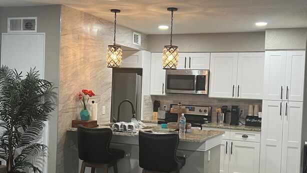 A modern kitchen with white cabinets, a central island with bar stools, a stainless steel microwave, and a built-in oven. - Luxe King Suite Bliss Getaway Condo Near Disney  (Kissimmee)