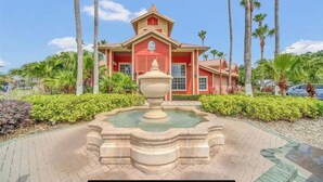 A red building with a fountain in front, surrounded by palm trees and greenery. - Luxe King Suite Bliss Getaway Condo Near Disney  (Kissimmee)