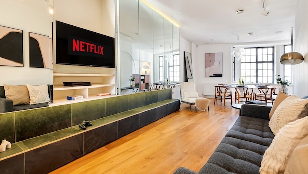 Ett modernt vardagsrum med en stor TV med platt skärm som visar ”NETFLIX”, en soffa, ett soffbord och en matplats med stolar och ett bord.