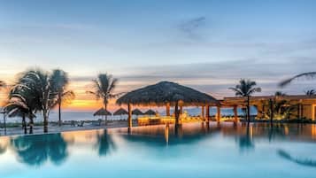 Un espace aménagé au bord de la piscine, offrant une vue sur le coucher du soleil, avec des palmiers et une structure dotée d’un toit de chaume.