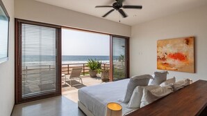 A bedroom with a large bed, a ceiling fan, a view of the ocean through floor-to-ceiling windows, and a painting on the wall. - Bright 2BR Villa I Sea Views + Open Kitchen (Zihuatanejo)