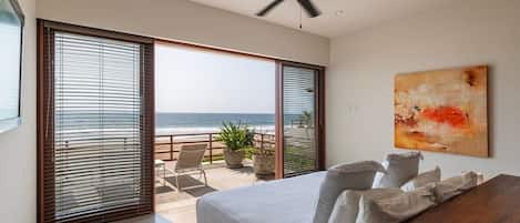 A bedroom with a large bed, a ceiling fan, a view of the ocean through floor-to-ceiling windows, and a painting on the wall.
