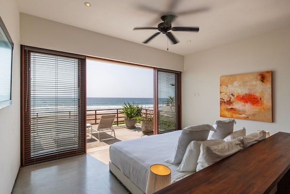 A bedroom with a large bed, a ceiling fan, a view of the ocean through floor-to-ceiling windows, and a painting on the wall.