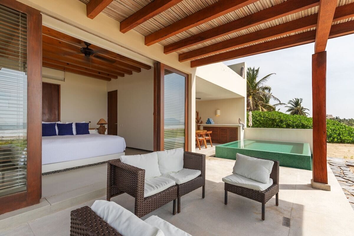 Une chambre avec un lit, deux fauteuils en osier, un ventilateur de plafond et une vue sur une piscine et la verdure.