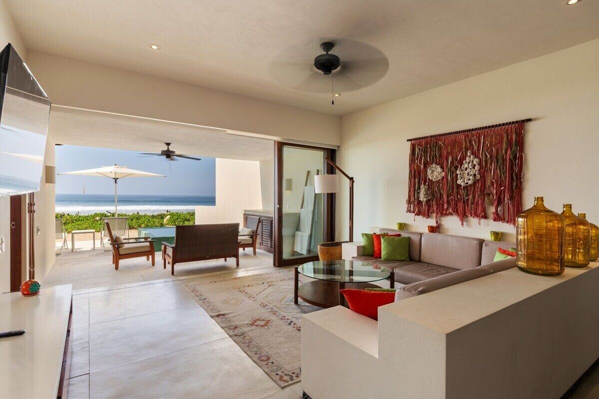 A modern living room with a glass coffee table, a sofa with cushions, a ceiling fan, and a view of the ocean through large windows.