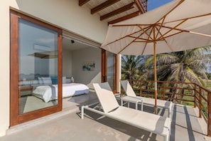A balcony with white lounge chairs and a large umbrella, overlooking a bedroom with a bed and a painting.