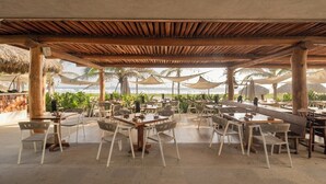 An outdoor dining area with tables and chairs, a wooden pergola overhead, and a view of the beach and palm trees. - 2BR Beachfront Villa + Pool, Style & Sea Views (Zihuatanejo)