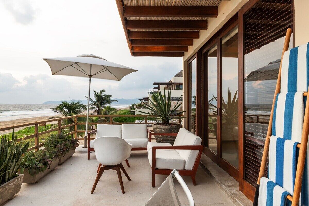 Varanda com vista para a praia, cadeiras estofadas brancas, um guarda-sol grande e plantas em vasos.
