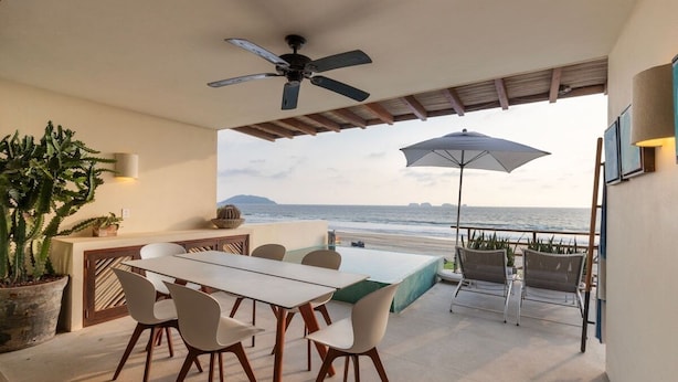 An outdoor patio with a dining table, chairs, a ceiling fan, a large umbrella, and a view of the ocean. - 2BR Beachfront Villa + Pool, Style & Sea Views (Zihuatanejo)