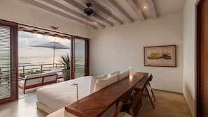 A bedroom with a bed, a wooden desk, and a view of the sea through large windows. - 2BR Beachfront Villa + Pool, Style & Sea Views (Zihuatanejo)