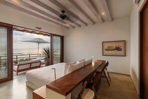 A bedroom with a bed, a wooden desk, and a view of the sea through large windows. - 2BR Beachfront Villa + Pool, Style & Sea Views (Zihuatanejo)