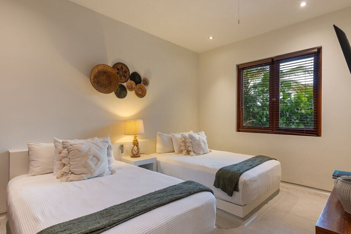 Habitación de hotel con dos camas, un adorno en la pared, una ventana con persianas y una mesita de noche con lámpara.