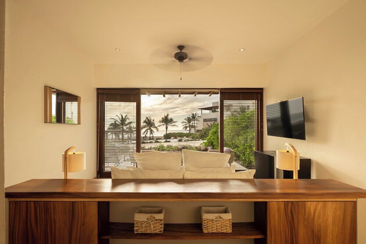 A modern hotel room with a wooden desk, a sofa, a TV mounted on the wall, and a view of palm trees and a beach.