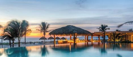 A poolside area with a sunset view, palm trees, and a thatched-roof structure.
