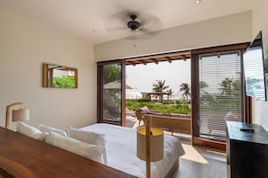 A hotel room with a bed, a desk, a chair, a lamp, a ceiling fan, and a large window with blinds. - Zihuatanejo 3BR • Beachfront Villa w/ Pool & Patio (Zihuatanejo)