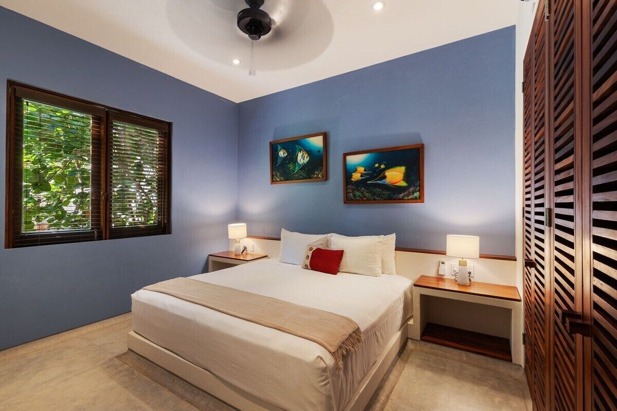 Une chambre à coucher avec un lit, deux tables de chevet, un ventilateur de plafond et des œuvres d’art accrochées au mur.