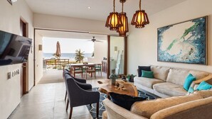 A modern living room with a sofa, chairs, a coffee table, and a TV. There is a balcony with a view of the ocean. - Elegant 2BR Villa I Ocean View + Artful Style (Zihuatanejo)