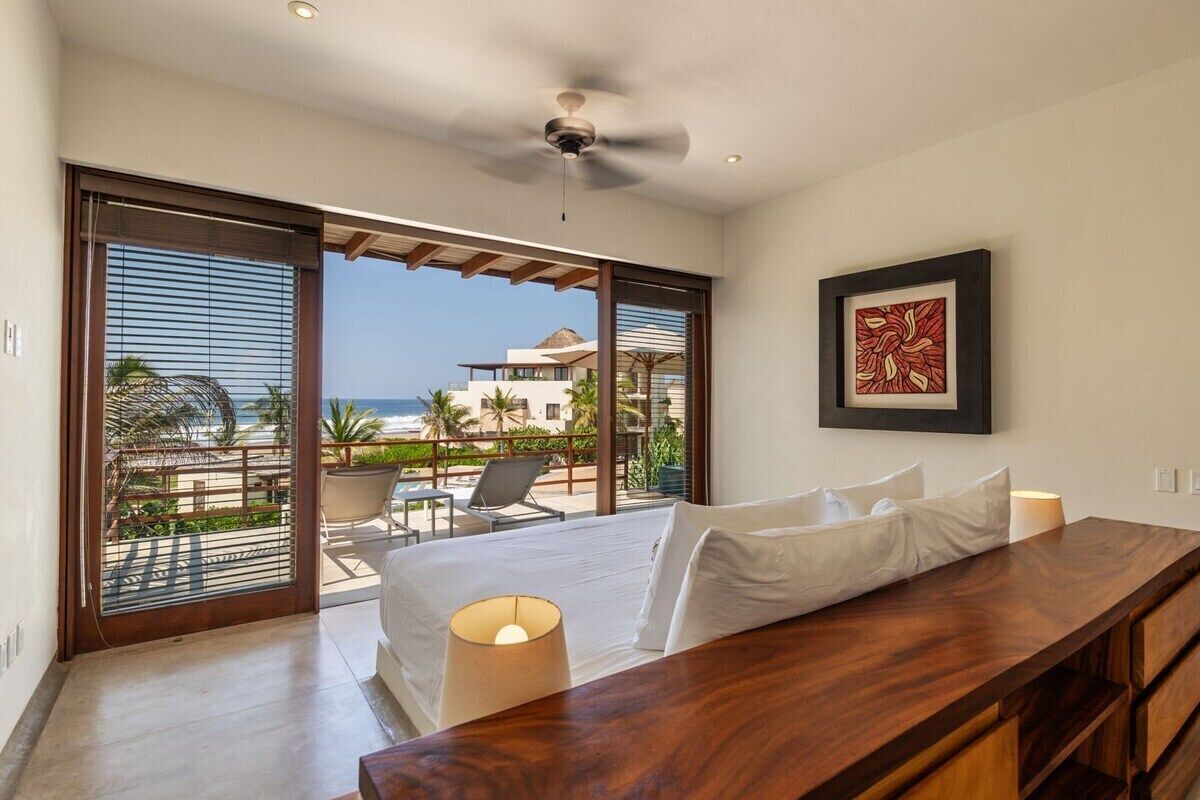 A hotel room with a bed, a wooden desk, a ceiling fan, a large window with a view of the beach, and a framed artwork on the wall.
