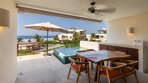 A balcony with a view of the ocean, a pool, and a dining area with wooden furniture. - 2BR Ocean View Apt | Exclusive Zihua Stay (Zihuatanejo)