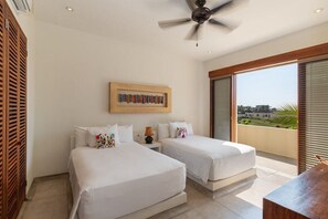 Habitación de hotel con dos camas, ventilador de techo, una obra de arte enmarcada sobre las camas y vista al exterior a través de amplias ventanas.