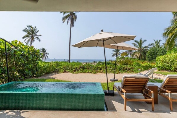 Zona de piscina con jacuzzi, sillas de descanso y sombrillas, con vistas a una playa con palmeras.