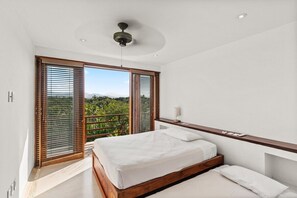 A bedroom with a bed, a bedside table, a ceiling fan, and a view of greenery through large windows. - Zihuatanejo 3BR Penthouse • Ocean Views + Dining (Zihuatanejo)