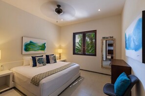 A hotel room with a bed, a desk, a chair, a window with blinds, and a ceiling fan. - Stylish 3BR Villa I Pool & Sunset Views (Zihuatanejo)