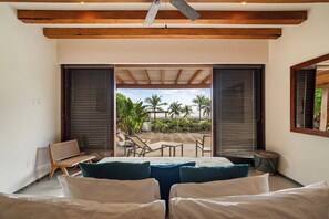 A bedroom with a bed, two windows with blinds, a ceiling fan, and a view of a beach with palm trees. - Stylish 3BR Villa I Pool & Sunset Views (Zihuatanejo)