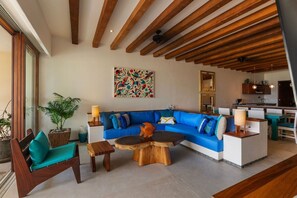 A modern living room with a blue sectional sofa, wooden coffee table, and a dining area with a kitchen in the background. - Stylish 3BR Villa I Pool & Sunset Views (Zihuatanejo)
