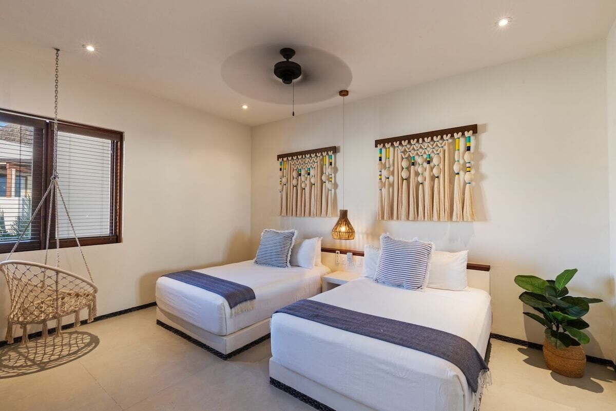 A hotel room with two beds, a hanging chair, a potted plant, and a window with blinds.