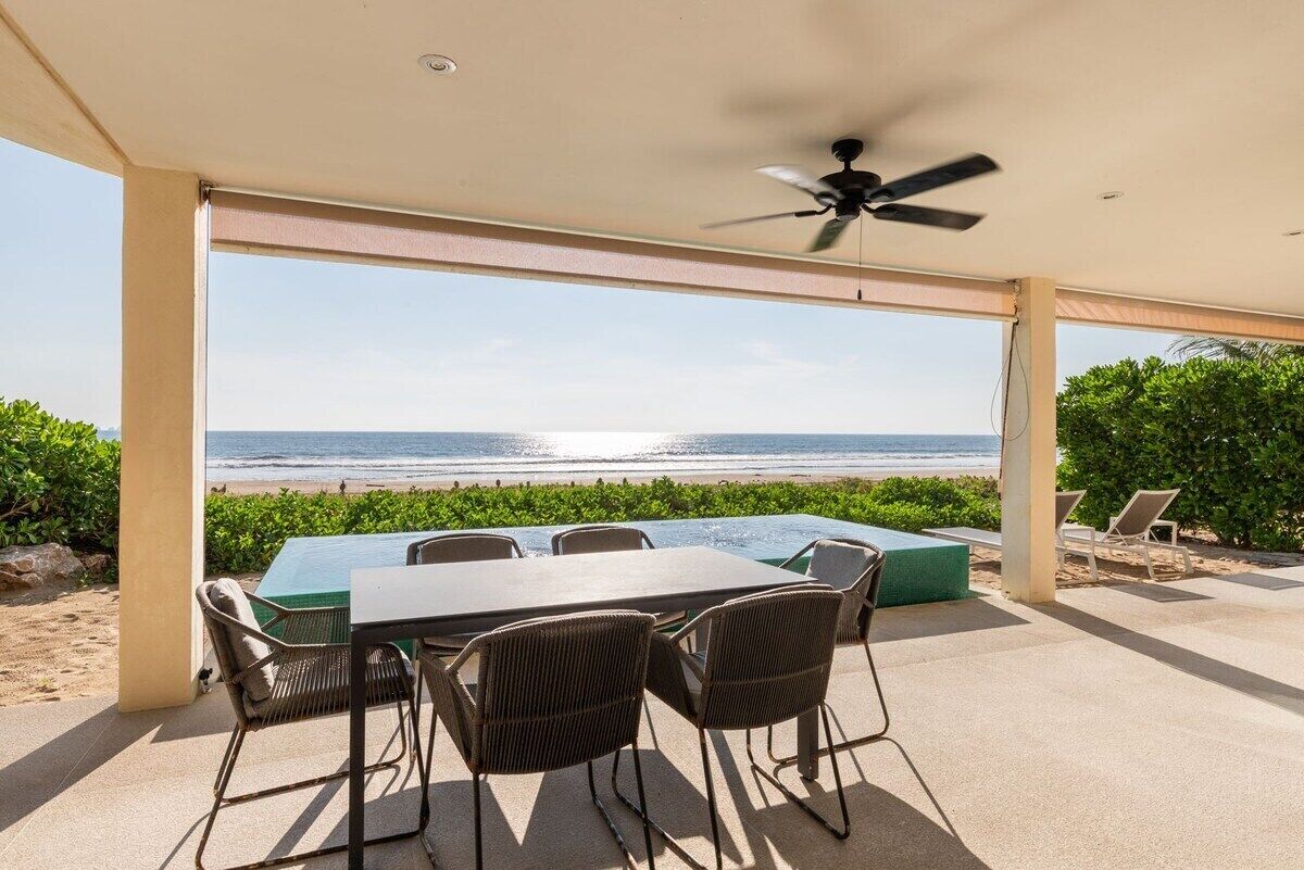 Een terras buiten met uitzicht op het strand, een tafel, stoelen en een plafondventilator.