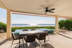An outdoor patio with a view of the beach, a table, chairs, and a ceiling fan.