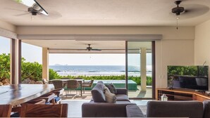 A modern living room with a large TV, a dining area, and a view of the ocean.