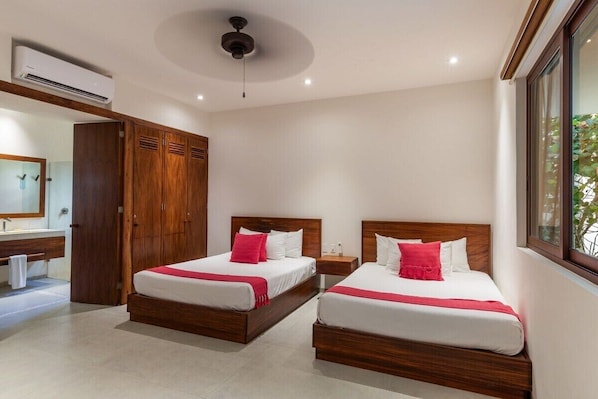 A hotel room with two beds, a wooden wardrobe, a bathroom with a mirror, and an air conditioning unit.