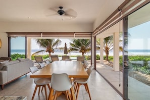 A modern dining area with a large table and chairs, overlooking a beach with palm trees.