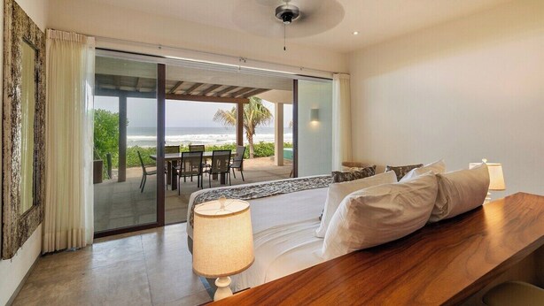 A bedroom with a large bed, a wooden nightstand, a lamp, and a view of the ocean through a sliding glass door. - 3BR Beachfront Villa • Private Terrace + Pool (Zihuatanejo)