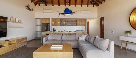 A modern living room with a large sectional sofa, a wooden coffee table, and a kitchen area with a hanging light fixture.