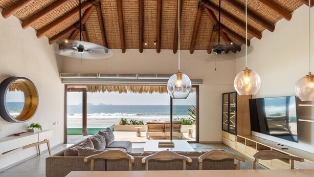 Ein modernes Wohnzimmer mit großem Fenster zum Strand, Deckenventilator und Flachbildfernseher.