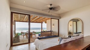 Ein Schlafzimmer mit einem großen Bett, einem Holztisch, einem Deckenventilator und Meerblick durch eine Schiebetür.