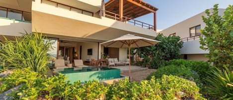 Un edificio moderno de dos pisos con piscina, mobiliario exterior y abundante vegetación.