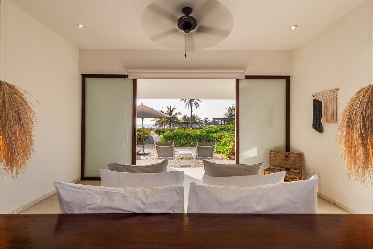 Quarto de hotel com cama, mesa de jantar e vista para uma praia com palmeiras.