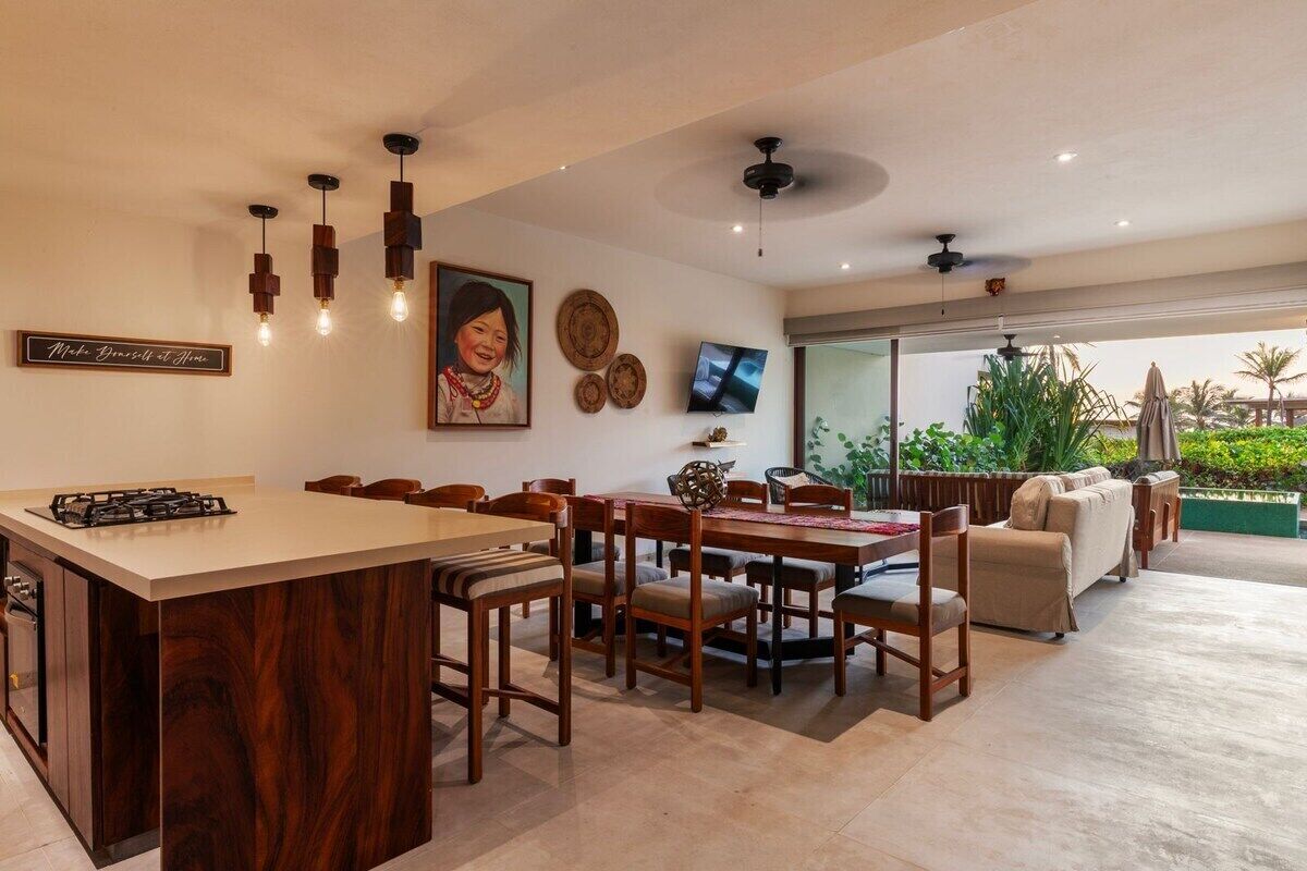 A modern kitchen with a central island, dining area with wooden chairs, a sofa, and a TV mounted on the wall.