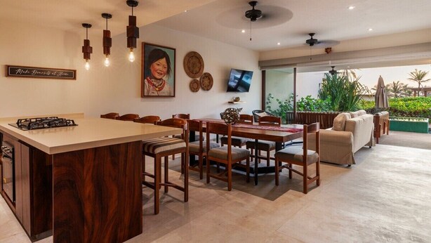A modern kitchen with a central island, dining area with wooden chairs, a sofa, and a TV mounted on the wall. - Zihua 3BR Villa • Oceanfront + Sleeps 6 + Family (Zihuatanejo)