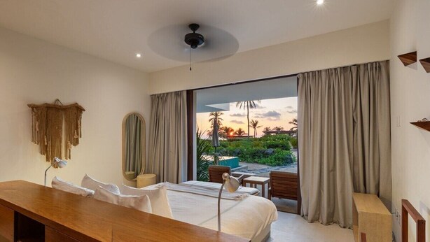 Una camera d'albergo con un letto, una scrivania, una sedia, una ventola a soffitto e vista su un giardino e un tramonto.