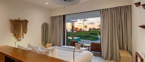 A hotel room with a bed, a desk, a chair, a ceiling fan, and a view of a garden and sunset.