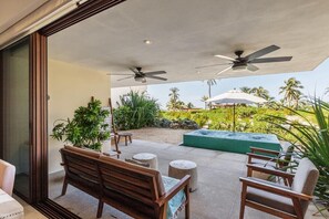 An outdoor patio with a wooden bench, two chairs, a small table, and a green planter. There are ceiling fans and a view of a landscaped garden with palm trees. - Family-Friendly 4BR I Sand at Your Door + Pool (Zihuatanejo)