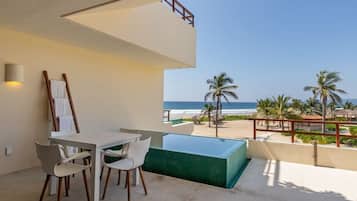 A balcony with a view of the ocean, a pool, and a dining area with chairs and a table.